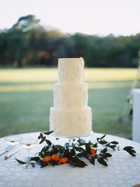Wedding cake, a four-tier cake with white-on-white floral detailing, styled with citrus fruit on a lawn table amid trees and open sky.