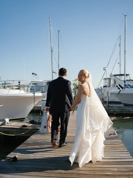 Bride and groom portrait on a wooden dock at a sunny marina, she holding a simple bouquet as sailboat masts rise against blue water and sky.