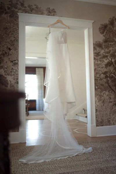 Wedding dress on hanger draped with a long bridal veil, framed in a doorway with wallpaper and a glimpse of the staircase.
