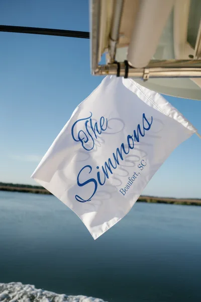 Custom wedding flag with blue lettering, set against a blue sky over water near a distant shoreline.