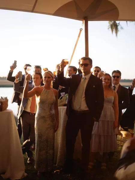 Wedding toast and reception cheers as guests raise cocktail glasses on a grass lawn by the waterfront, framed by a warm sunset sky.