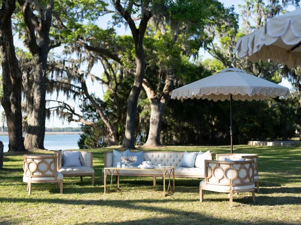 Wedding lounge setup for outdoor cocktail hour seating with a tufted sofa on a grass lawn beneath oak trees draped in Spanish moss by the water.