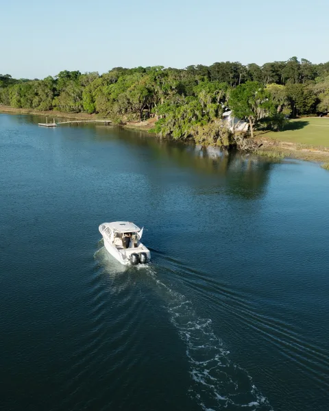 Wedding boat exit as a couple departing by boat aboard a motorboat along a calm river, past shoreline trees and a small dock under blue sky.