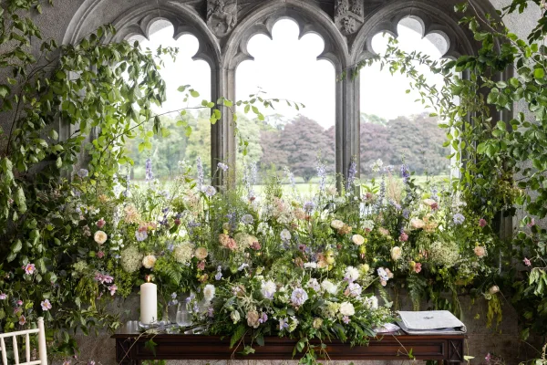 Wedding ceremony backdrop with floral ceremony backdrop meadow blooms and greenery garland framing stone arch windows, candles on a wooden table