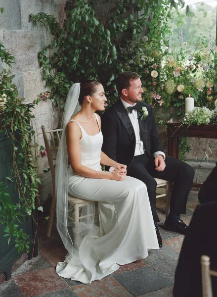 Ceremony moment as bride and groom seated holding hands, her cathedral veil draped beside wooden chairs on a stone garden patio