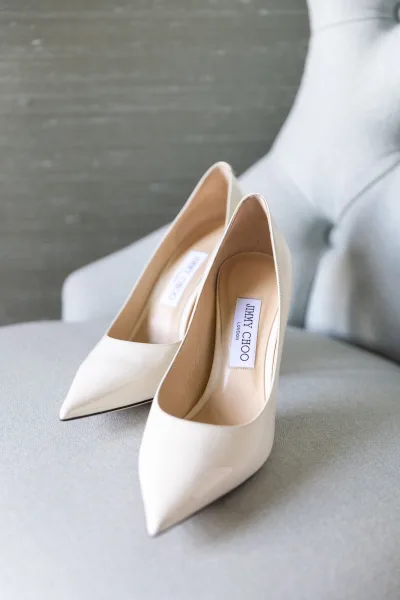 Bridal shoes, white wedding heels with pointed toes and a designer label resting on an upholstered chair against a gray wall