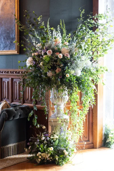 Ceremony floral arrangement with roses and cascading greenery in a pedestal urn, set against a wood paneled wall with window light