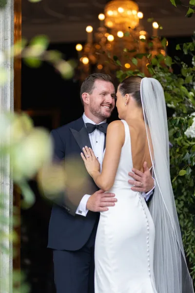 Couple portrait of bride and groom embrace, bride showing wedding ring, under chandelier warm lights in a doorway with greenery backdrop