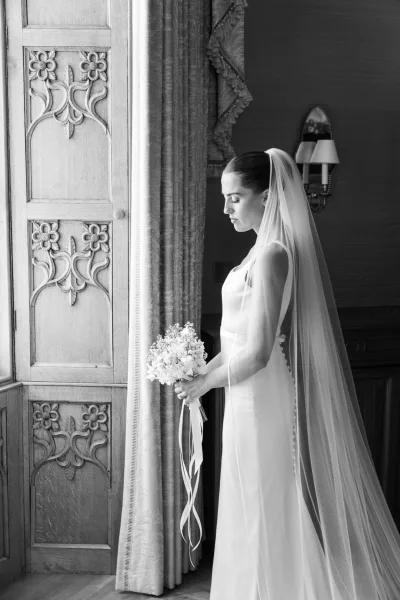 Bridal portrait in black and white, bride in profile by window light holding a bouquet, long veil draped near a carved wooden door