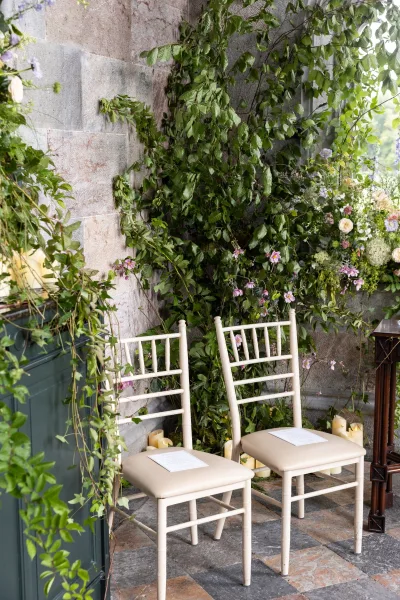 Ceremony seating with white chiavari chairs lined with greenery garland, pastel flowers, and pillar candles against a stone wall patio