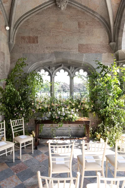 Ceremony setup with greenery wedding altar, floral installation and candles beside a table, white chiavari chairs in stone courtyard archway