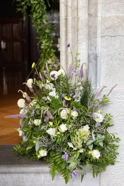 Ceremony floral arrangement of wedding ground florals with lush greenery and white and purple blooms on stone steps by a wooden doorway