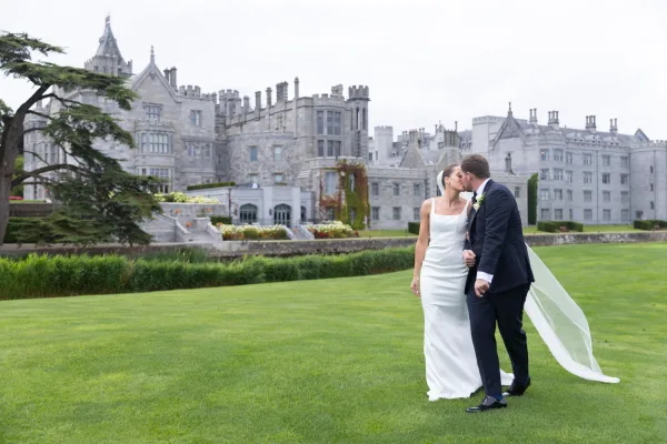 Wedding kiss as bride in a minimal satin wedding dress and long veil kisses groom in navy suit before a castle garden lawn under overcast sky