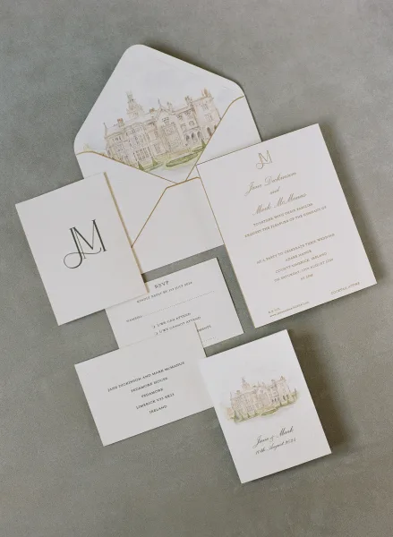 Invitation suite with gold script cards, monogram and RSVP details, plus a gold-lined envelope and watercolor venue illustration on gray backdrop