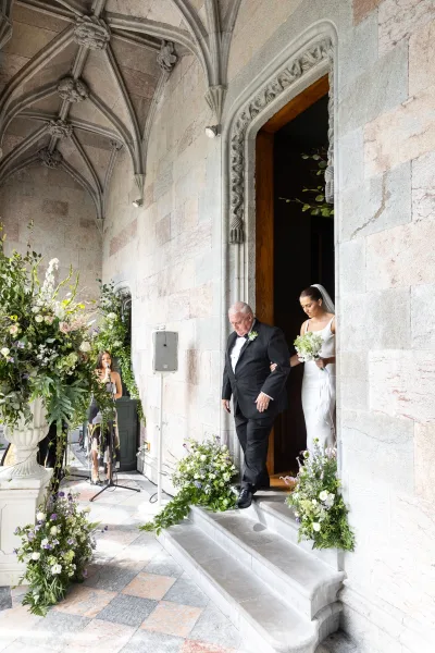 Wedding processional as father walks bride in satin gown and veil holding bouquet through stone church archway with urn florals on steps