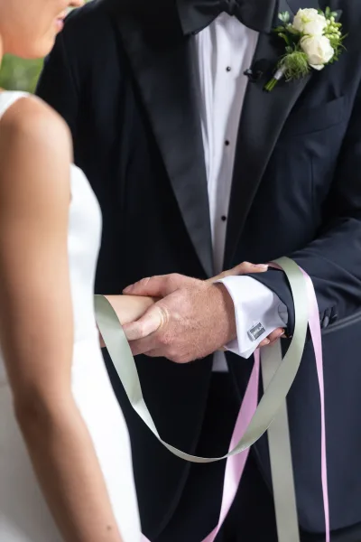 Ceremony handfasting as bride and groom clasp hands tied with satin ribbons, groom in black tuxedo and white rose boutonniere in blurred garden