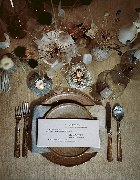 Reception tablescape with wedding place setting, menu card on stoneware plates, linen napkin, amber goblets, dried florals, bird nest with speckled eggs on linen tablecloth