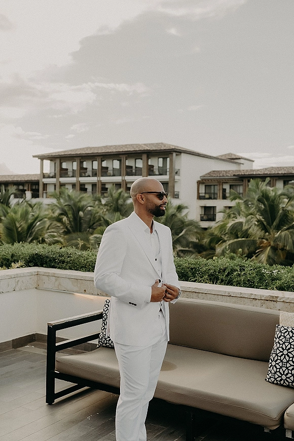 Groom portrait in a white suit wearing sunglasses on a terrace patio lounge sofa with palm trees and a resort building behind under cloudy sky