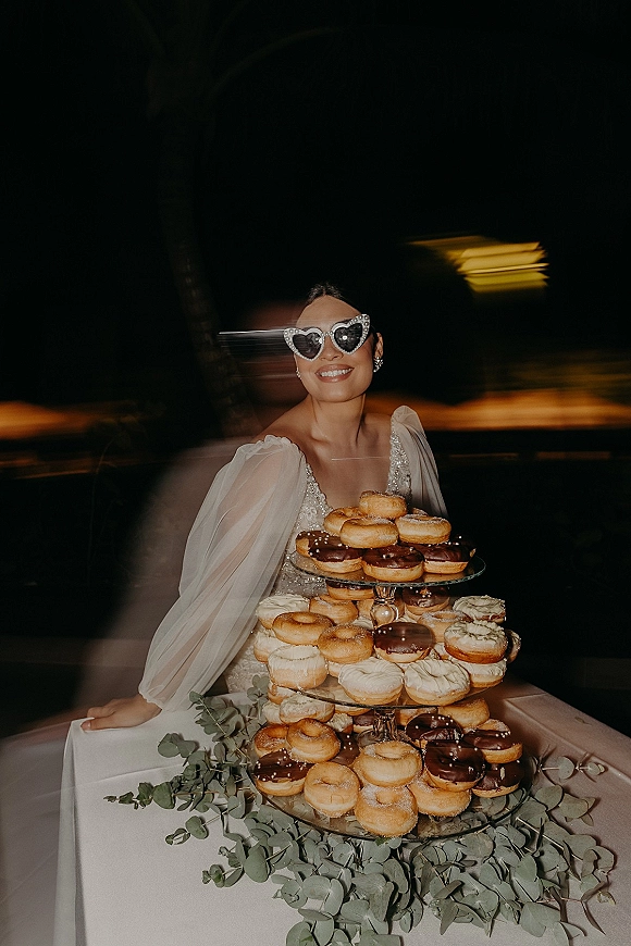 Bride portrait in heart sunglasses beside a donut tower dessert stand, sparkly wedding dress and earrings against blurred night lights