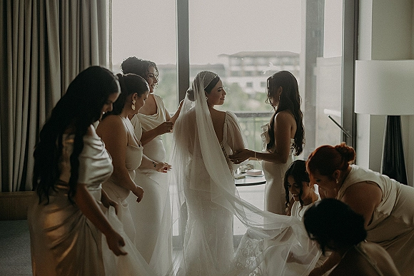 Bride getting ready as bridesmaids help adjust her bridal gown and wedding veil by a large hotel room window overlooking city buildings