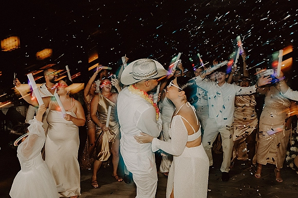 Wedding reception dancing as bride in gown and glasses and groom in white suit and cowboy hat twirl amid confetti, glow sticks on deck at night