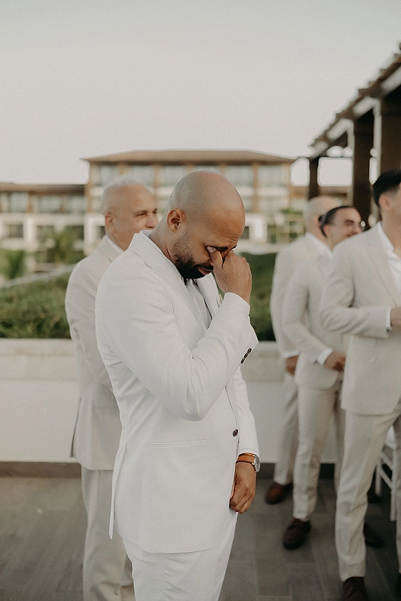 Groom portrait of a man in a white suit crying and wiping tears, boutonniere and watch visible, on an outdoor terrace with groomsmen behind
