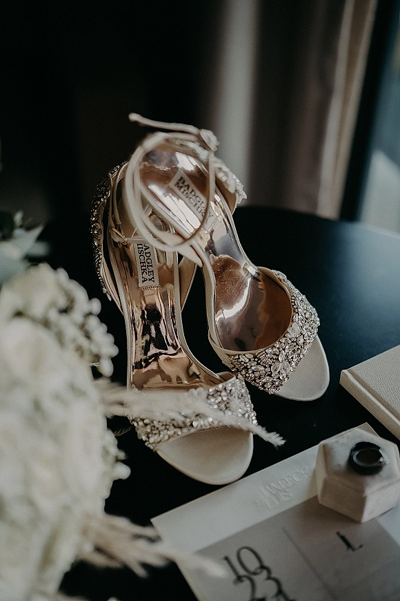 Wedding shoes and bridal heels with rhinestone accents, ankle straps, and open toes beside invitation suite and ring box on dark tabletop by window light