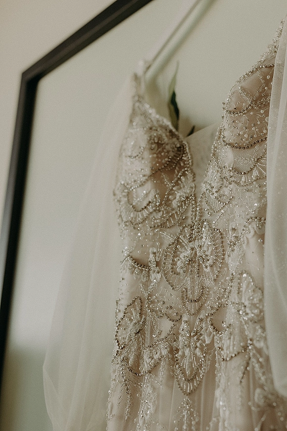 Wedding dress close-up showing beaded wedding dress bodice with lace appliqué and sheer tulle, hanging on a hanger against a light wall.