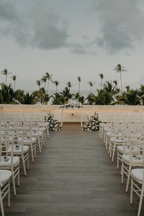 Ceremony setup for an outdoor wedding ceremony with white chiavari chairs and a candle-lined aisle runner facing an oceanfront floral arch on a terrace