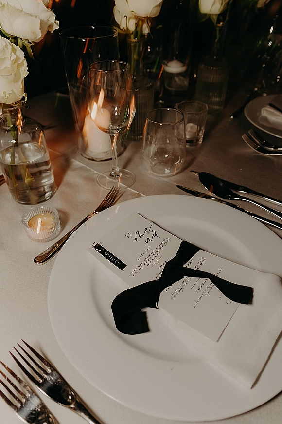 Wedding place setting with wedding menu card tied in a black ribbon on a white plate, surrounded by glassware, roses, and candlelight