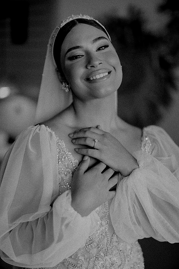 Bridal portrait of a smiling bride in a wedding veil, showing her ring and beaded dress with pearl earrings against a blurred indoor background