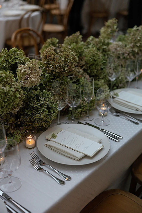 Wedding tablescape with hydrangea garland centerpiece, white table settings, menu cards, and votive candles on round tables in an indoor reception space