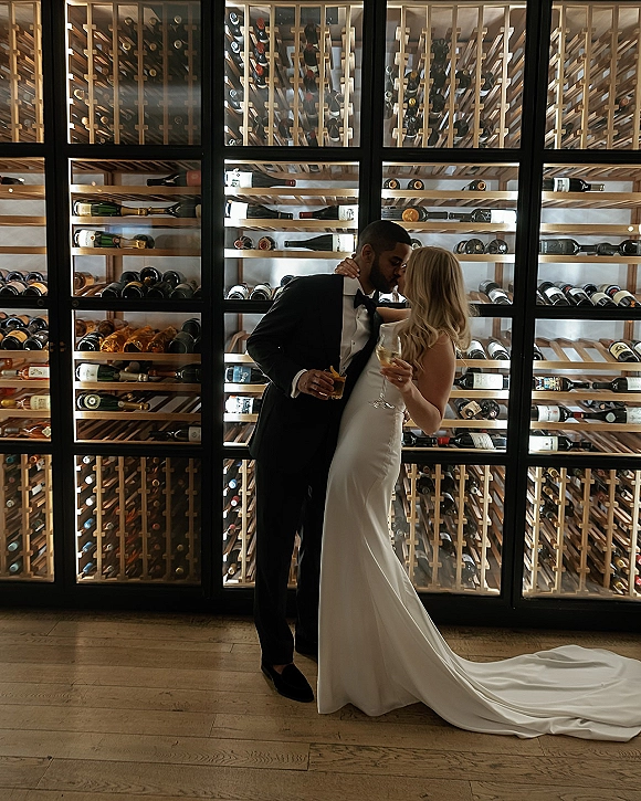 Wedding kiss portrait of bride and groom kissing, holding wine glasses in a wine cellar with glass wall wine bottles behind them