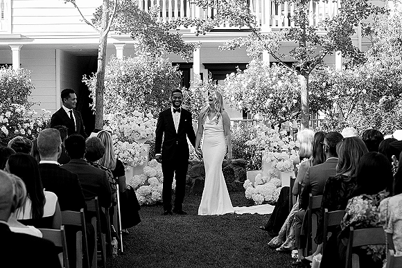 Ceremony moment at an outdoor wedding ceremony with bride and groom holding hands by a floral arch on a garden lawn with guests seated