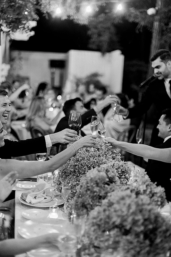 Wedding reception toast with champagne flutes raised as guests clink glasses around a long table on an outdoor patio under string lights