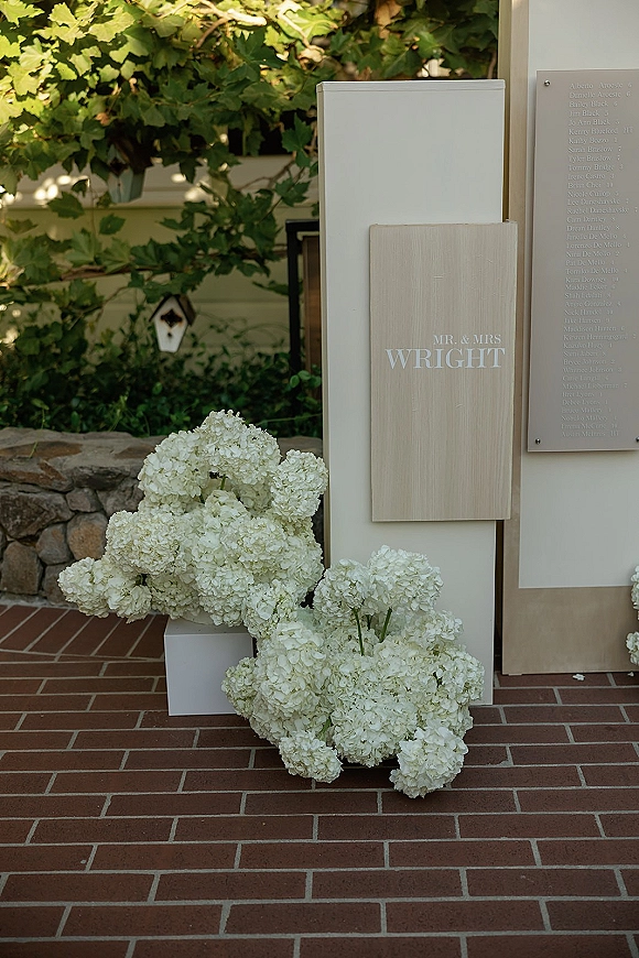 Wedding welcome sign on a white pedestal with white hydrangeas, next to an acrylic seating chart along a brick walkway by a stone wall