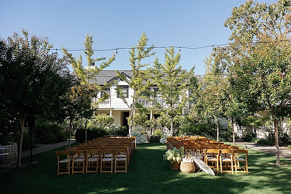 Outdoor ceremony setup with backyard wedding ceremony seating, wood chairs lining a grass aisle with string lights and white florals by a house