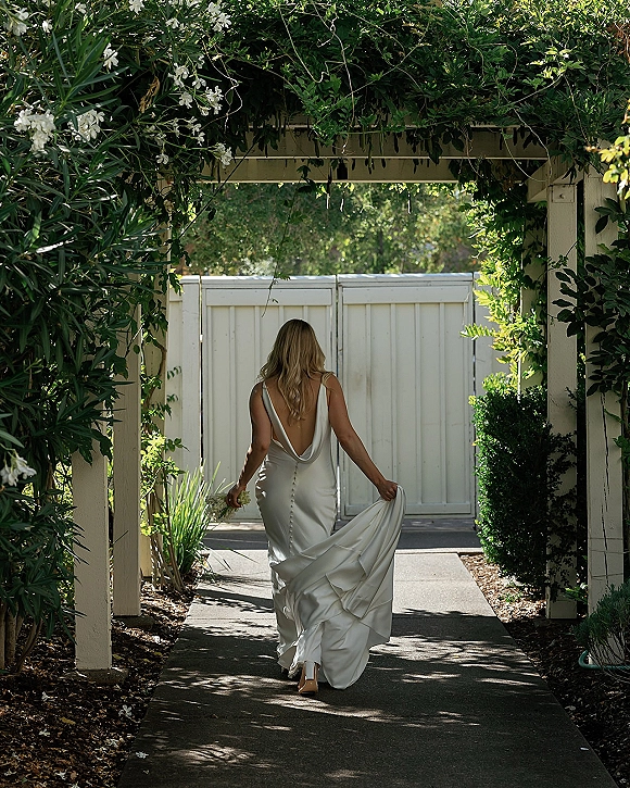 Bridal portrait of a bride walking away in a backless wedding dress, holding her train and bouquet on a sunlit garden pergola walkway