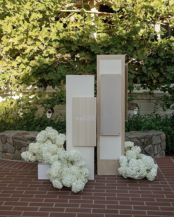 Wedding welcome sign with modern wedding welcome sign design, framed by white hydrangeas on a brick patio near a vine-covered stone wall