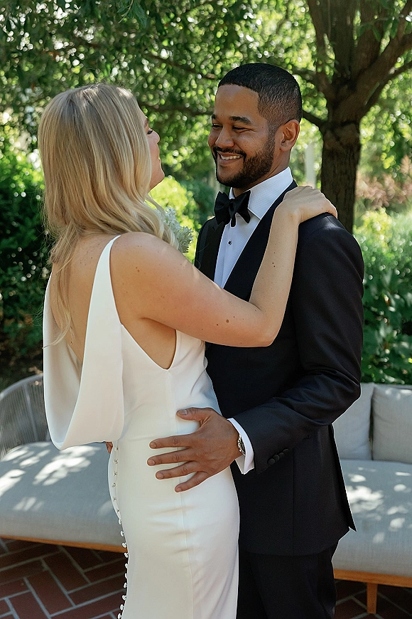 Couple portrait of bride and groom embrace, bride in white dress hugging groom in black tuxedo on a garden patio with greenery
