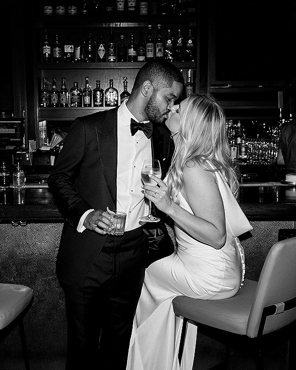 Wedding kiss portrait of bride in strapless dress and veil kissing groom in black tux at bar, holding wine and whiskey glasses