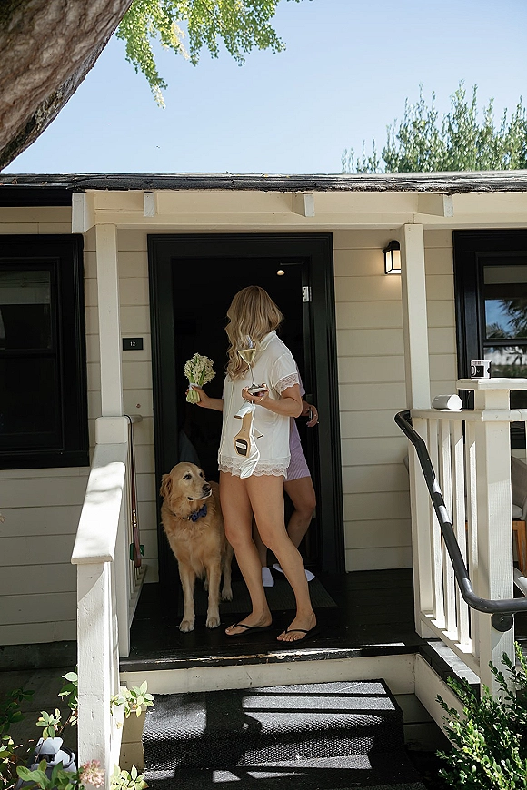 Getting ready moment as bride in robe holds a white bouquet and heels, sipping champagne on porch steps with dog in bow tie, blue sky behind