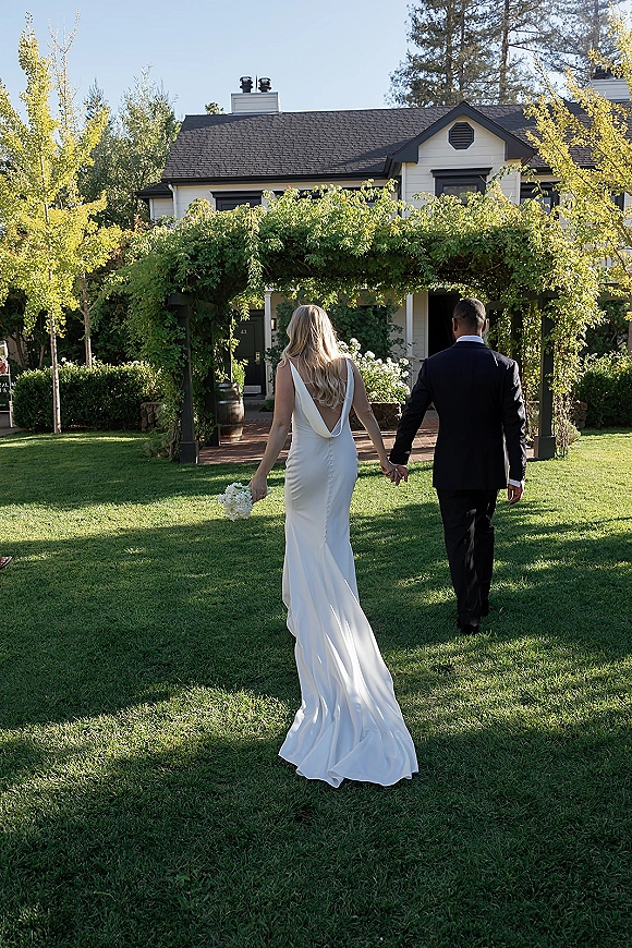Couple portrait of bride and groom walking away holding hands under a greenery arch, her bouquet and veil trailing on a garden lawn