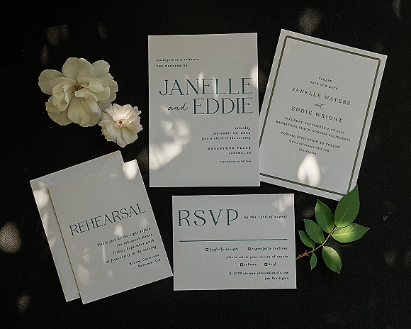 Invitation suite with wedding invitation suite cards, envelopes, and white flowers with greenery arranged on black fabric in dappled sun