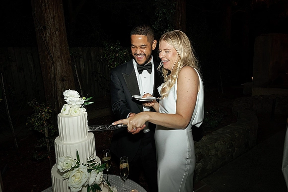 Wedding cake cutting as bride and groom slice a two-tier white cake with rose flowers and greenery on a dessert table in a garden at night