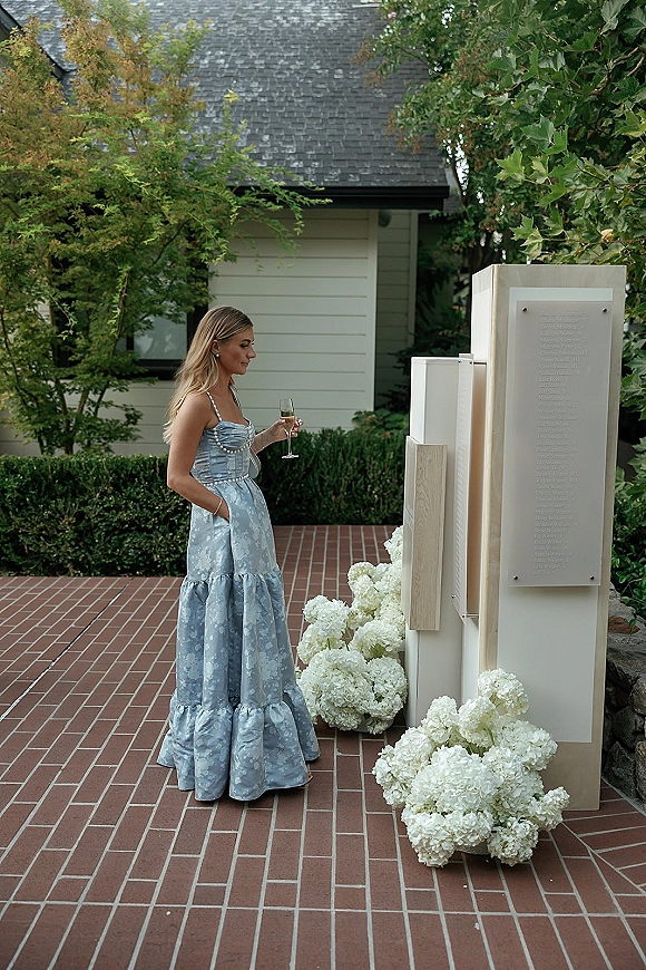 Wedding guest portrait in a strapless blue dress holding a champagne flute beside white hydrangeas and modern signage on a brick patio