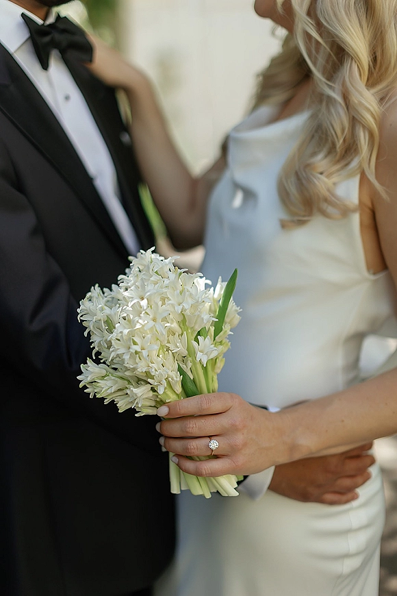 Couple portrait of bride holding bouquet with her engagement ring visible as groom in black tuxedo embraces her against blurred garden greenery