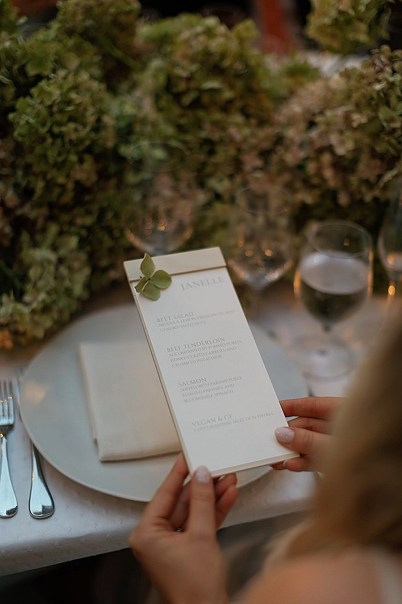Wedding menu card with clean wedding menu design on a dinner plate, finished with a greenery sprig at an elegant reception table setting