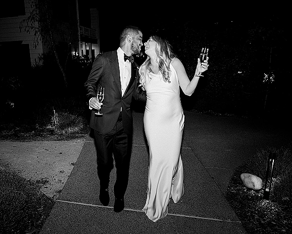 Wedding couple portrait of bride and groom walking with champagne flutes, laughing under outdoor lights along a garden path at night