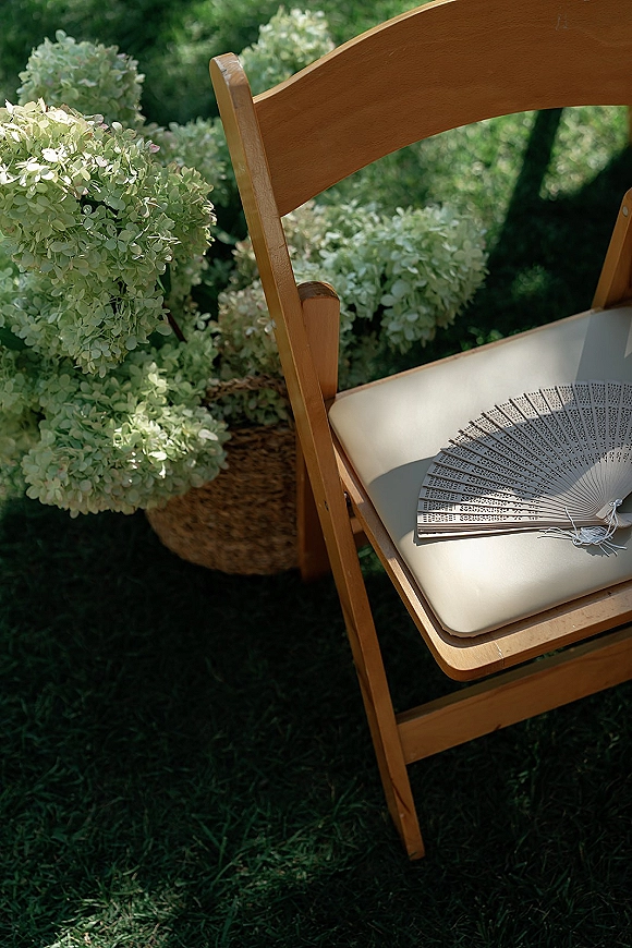 Ceremony chair decor with a paper hand fan on a cushioned wood folding chair, beside a woven basket of white hydrangeas on grass lawn